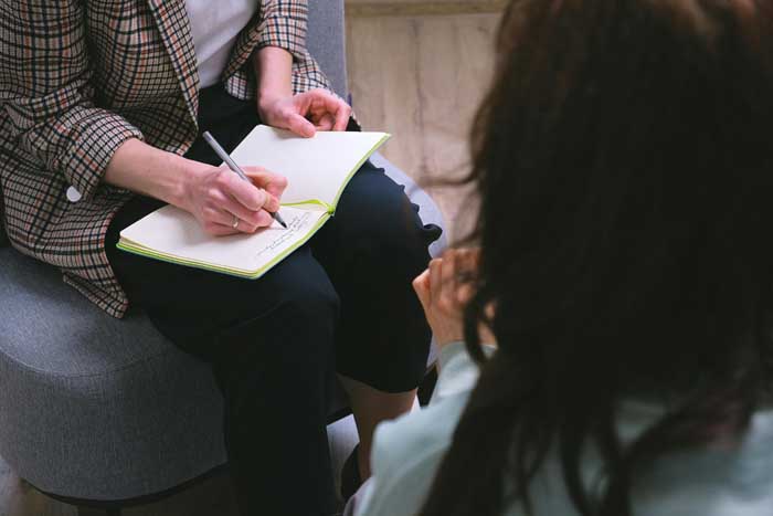 Eine Frau sitzt in der Psychosozialen Beratung. Beraterin notiert im Notizbuch mit.